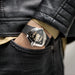 Close-up of a person wearing a black leather watch with a detailed face, set against a dark background.