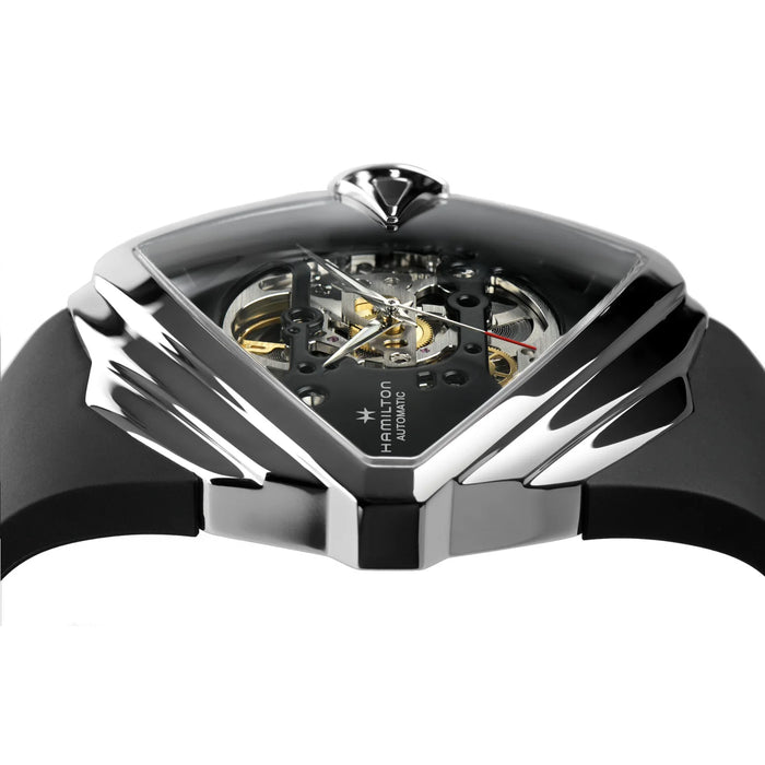 A close-up of a luxury skeleton watch with a black case, transparent dial showing the inner gold and silver mechanical movement, and a black strap on a white background.