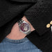 Hand wearing a silver watch with a dark face, against a black coat background