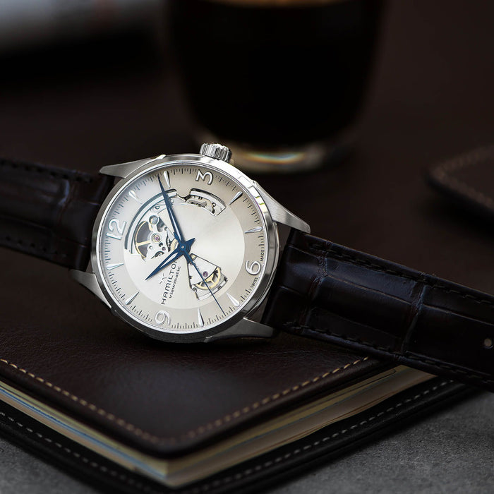 A luxury wristwatch with a silver-toned case, skeleton dial, and blue hands, resting on a dark brown leather notebook with a blurred dark beverage in the background.