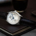 A luxury wristwatch with a silver-toned case, skeleton dial, and blue hands, resting on a dark brown leather notebook with a blurred dark beverage in the background.