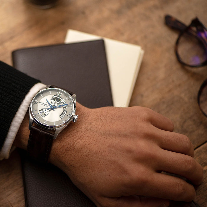 Hand wearing a watch on a wooden surface with a notebook and glasses.