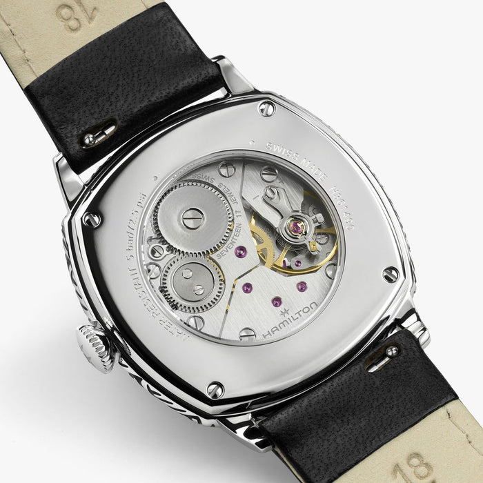 Close-up of the back of a Hamilton watch with a silver-toned case, transparent screw-down back, visible mechanical movement, and a black leather strap on a light gray background.