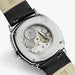 Close-up of the back of a Hamilton watch with a silver-toned case, transparent screw-down back, visible mechanical movement, and a black leather strap on a light gray background.
