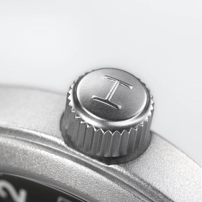 Close-up of a silver metal watch crown button engraved with the letter T, set against a clean light gray background.