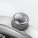 Close-up of a silver metal watch crown button engraved with the letter T, set against a clean light gray background.