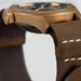 Close-up of a bronze watch with a brown leather strap on a light gray background