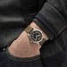 Hand wearing a watch with a dark face and brown leather strap, against a dark background.