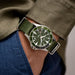 Green watch with a green strap on a person's wrist, wearing a green jacket and blue pants.