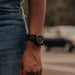 A close-up lifestyle shot of a person wearing a minimalist black wristwatch with a black strap and gold-tone hands, paired with blue denim jeans.